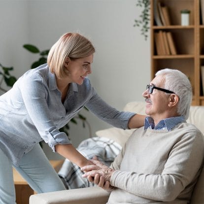 A home carer leaning in to hold hands and reassure a smiling older man in his living room