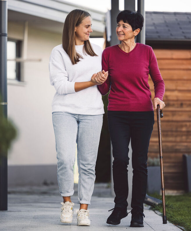 A smiling caregiver walking arm-in-arm with an older woman using a cane outside her home, representing compassionate, person-centred support.