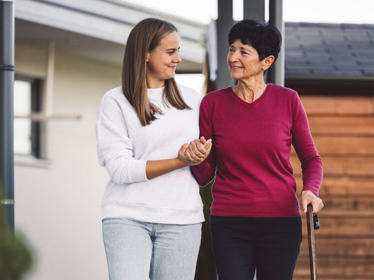 A smiling caregiver walking arm-in-arm with an older woman using a cane outside her home, representing compassionate, person-centred support.