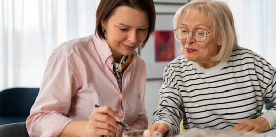 A caregiver and an older woman smiling together while reviewing a notebook at a table, representing collaborative care.