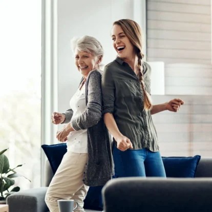 A caregiver and an older woman laughing joyfully while dancing together in a bright living room, showcasing emotional support and companionship.