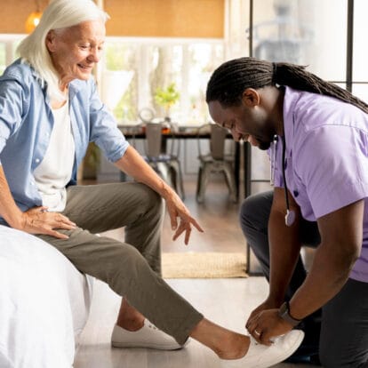 A smiling male caregiver kneeling to cheerfully help an older woman tie her shoes as she sits on her bed, illustrating respectful assistance with dressing.