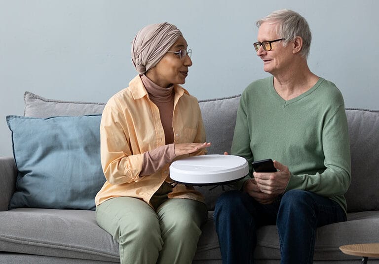 A female caregiver and an older man sitting comfortably on a living room sofa, smiling as they look at a smart home device and a smartphone together.
