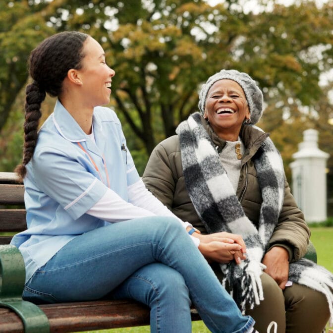 A female caregiver in uniform and an older woman sitting together on a park bench, holding hands and laughing joyfully, representing safe, trusted, and compassionate care.